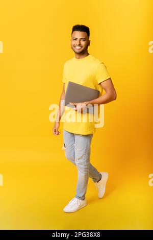 Allegro indiano uomo che porta computer portatile isolato su giallo. Allievo arabo maschio felice con il laptop che cammina, freelancer usando il gadget portativo, il calcolatore. Foto verticale Foto Stock