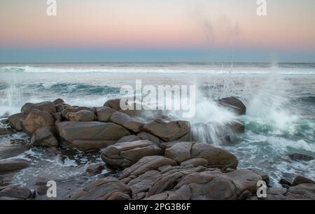 Scena costiera che mostra un'onda che si schiantano sulle rocce con una grande onda che si interrompe in lontananza Foto Stock