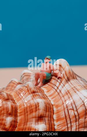 primo piano di un uomo in miniatura, in costume da bagno verde, con cappuccio verde e un paio di occhiali da sole, seduti su una conchiglia, su sfondo blu con s Foto Stock