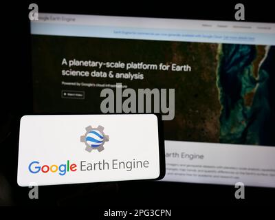 Persona che tiene lo smartphone con il logo del prodotto software Google Earth Engine sullo schermo davanti al sito Web. Messa a fuoco sul display del telefono. Foto Stock
