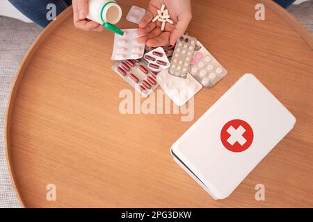 Vista dall'alto di un maschio senza volto di raccolto che prende compresse da una bottiglia di plastica con vari rifornimenti di medicina posto vicino alla scatola di primo soccorso su un tavolo di legno a casa Foto Stock