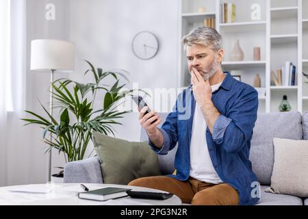 Uomo adulto maturo rovesciato e confuso leggendo le notizie difettose in linea dal telefono, persona grigia haired che siede sul divano nel soggiorno nel paese che lavora con le finanze domestiche dei documenti ed il preventivo sul lavoro di carta. Foto Stock