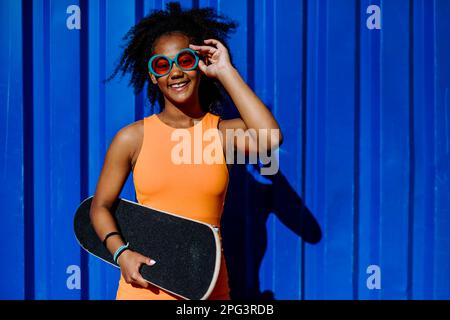 Ritratto di ragazza adolescente multirazziale con skateboard e occhiali da sole, di fronte al muro blu durante il giorno estivo. Foto Stock