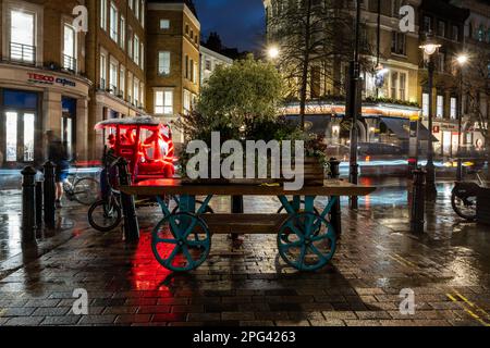 Un tradizionale carrello a mano decorato con scatole di piantatrice in legno blocca i veicoli a motore da King Street vicino a Covent Garden nel centro di Londra. Foto Stock