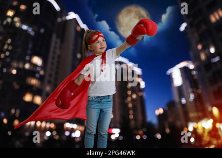 Bambina con costume da supereroe e splendido paesaggio urbano di notte sullo sfondo Foto Stock