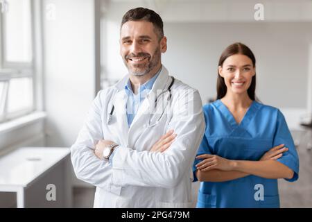 Felice medico e colleghi di infermiera femminile, indossando cappotti, in piedi con le braccia piegate, in posa in ufficio medico Foto Stock