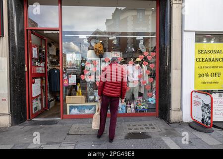 Un uomo che indossa una giacca rossa guarda all'esposizione frontale di una vetrina di beneficenza, situata su Putney High Street, a sud-ovest di Londra, Inghilterra, Regno Unito Foto Stock