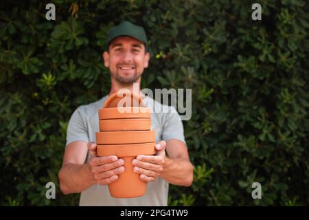 Primo piano ritratto uomo giardiniere in berretto verde tiene in mano pentole di terracotta terracotta contro lo sfondo di un recinto sempreverde. Giardinaggio, piccola impresa, Foto Stock