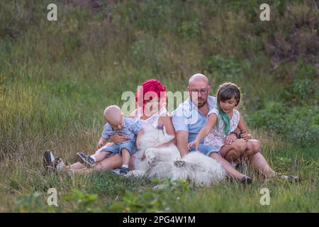 Ritratto di famiglia luminosa, insolita, diversità in natura con il cane Samoyed. Viaggiare con animali domestici. Le persone creative guardano la fotocamera. Verde botanico bac Foto Stock