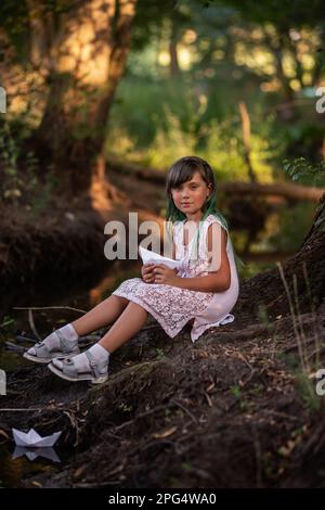 Ritratto di ragazza con capelli verdi in vintage abito, siede sulla riva del fiume, tenendo in mano bianco origami carta barca. Bambino lancia allegramente la sh Foto Stock