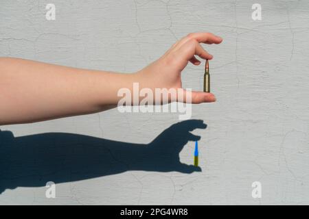 Cartuccia del fucile in mano. Le forme dell'ombra della mano sul muro. L'ombra della cartuccia è di colore giallo-blu. Concetto di sostegno militare per l'Ucraina Foto Stock