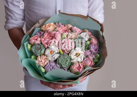 L'uomo senza volto tiene in mano un bouquet di fiori dolci di marshmallow. Confezione regalo in verde pastello con delicate rose senza zucchero su fondo grigio. Piccola Foto Stock