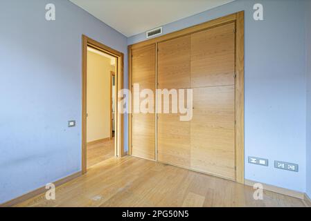 Una stanza vuota con un pavimento in legno di quercia chiaro, un armadio a muro con porte a battente in legno abbinate, una finestra con tende e pareti blu Foto Stock