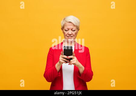 Donna bionda anziana con capelli corti che guarda lo schermo dello smartphone mentre si parla o si comunica in chat video su sfondo giallo Foto Stock