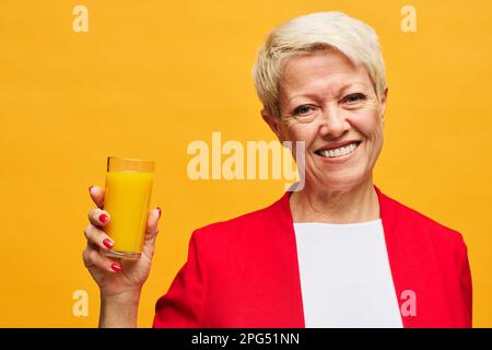 Allegra donna bionda matura con un sorriso toothy che tiene un bicchiere di succo d'arancia e guarda la macchina fotografica mentre si trova su sfondo giallo Foto Stock
