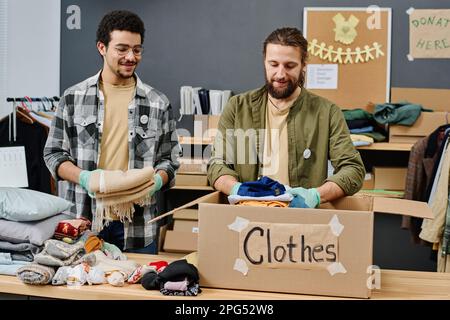 Due giovani volontari maschi interculturali felici preparano abiti di seconda mano per le persone bisognose e li mettono in scatola di cartone Foto Stock