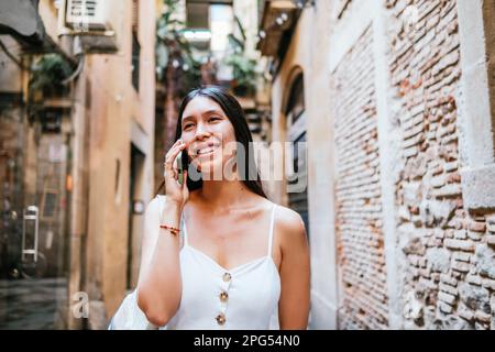 Felice donna latino-americana guardando via con sorriso e parlando su smartphone vicino a muro di mattoni di vecchio edificio sulla strada stretta di Barcellona, Spagna Foto Stock