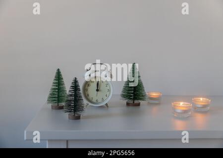 Una sveglia bianca, un puntatore a dodici, un piccolo albero di Natale artificiale verde e candele accese in candelabri su una cassettiera bianca. Copia Foto Stock