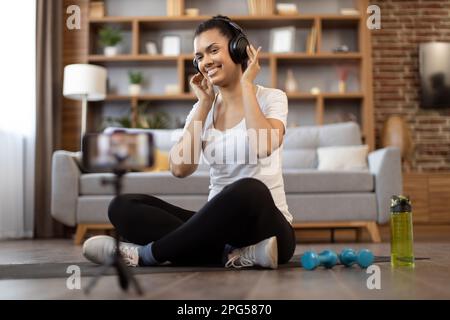 Grazioso vlogger multirazziale in cuffie sorridente alla fotocamera del telefono sul treppiede mentre si siede sul tappetino yoga vicino a manubri e bottiglia d'acqua. Donna attiva che registra video di allenamento per sport vlog in appartamento. Foto Stock