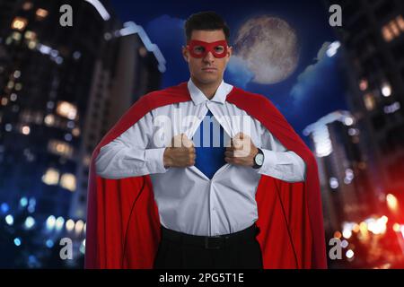 Uomo d'affari che indossa un costume da supereroe e un bellissimo paesaggio urbano di notte sullo sfondo Foto Stock