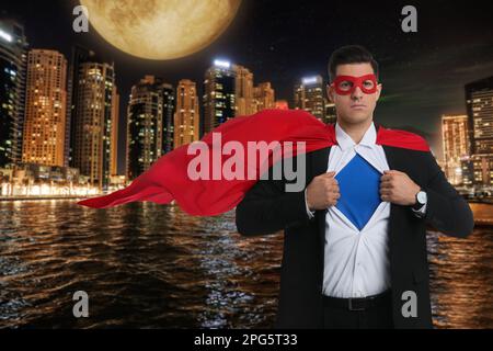 Uomo d'affari che indossa un costume da supereroe e un bellissimo paesaggio urbano di notte sullo sfondo Foto Stock