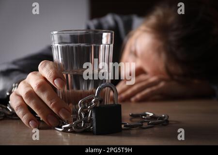 Dipendenza da alcol. Donna incatenata con un bicchiere di vodka che dorme su un tavolo di legno in camera Foto Stock