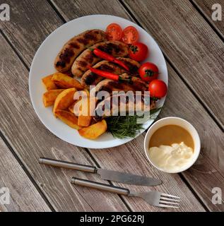 Un piatto con diverse salsicce alla griglia, patate, pomodori ciliegini, peperoni e erbe, posate e una tazza di salsa nelle vicinanze. Vista dall'alto, piatta Foto Stock