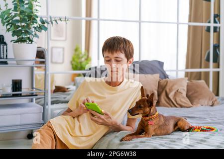Donna matura con un telefono in mano con un pygmy pinscher in camera da letto trascorre il tempo a casa godendosi il tempo libero, scattando foto e pubblicando sui social network Foto Stock