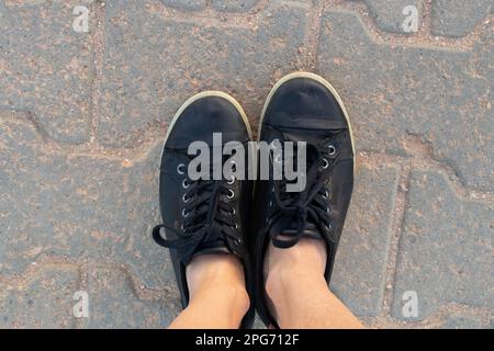 le gambe femminili in sneaker nere d'estate si alzano in piedi sulla strada una vista dall'alto Foto Stock