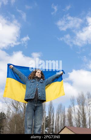 La giovane donna, una ragazza adulta, tiene una bandiera Ucraina giallo-blu dietro la schiena contro il cielo. Gli ucraini sono contrari alla guerra. Attirare l'attenzione su m Foto Stock