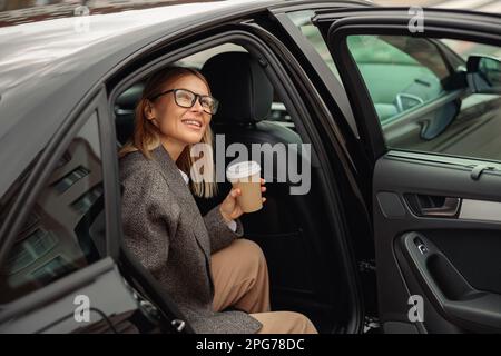 Donna elegante sorridente che indossa occhiali con caffè da asporto che esce dall'auto Foto Stock