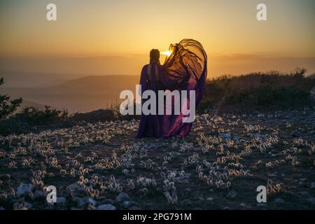 Tramonto viola abito donna montagne. Ascesa del mistico. tramonto sulle nuvole con una ragazza in un lungo vestito viola. Nel prato c'è un prato Foto Stock