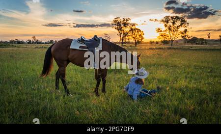 La ragazza si siede accanto al cavallo in campo aperto al tramonto Foto Stock