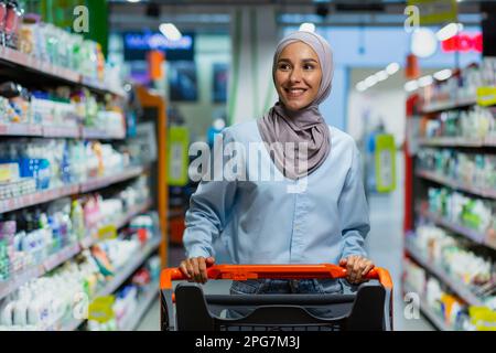 Ritratto di successo soddisfatto donna acquirente in supermercato, allegra donna musulmana in hijab con carrello per la spesa tra gli scaffali con merci che guardano vari negozi. Foto Stock