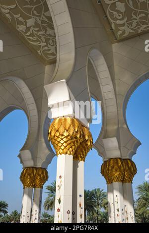 Vista del portico ombreggiato a piedi con colonne sormontate da un tronco di palma d'oro dipinto. Alla Grande Moschea dello Sceicco Zayed ad Abu Dhabi, Emirati Arabi Uniti, Foto Stock