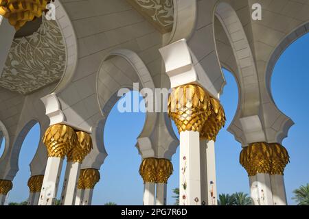 Vista del portico ombreggiato a piedi con colonne sormontate da un tronco di palma d'oro dipinto. Alla Grande Moschea dello Sceicco Zayed ad Abu Dhabi, Emirati Arabi Uniti, Foto Stock