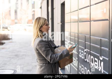 Bella donna che raccoglie un pacco da un elegante cassetta di sicurezza elettronica in acciaio, cassette postali automatiche. Servizio di consegna Paczkomat, ritiro Foto Stock