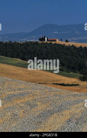 Stoppia di grano arato in terra argillosa e arancio nelle Crete Senesi contrasta con boschi, praterie e scintillanti colline azzurre in questa vista della Cappella della Madonna di Vitaleta in Val d’Orcia, Toscana, Italia. La cappella, fondata prima del 1590, è un simbolo iconico del Rinascimento italiano, patrimonio dell'umanità dell'UNESCO. Foto Stock