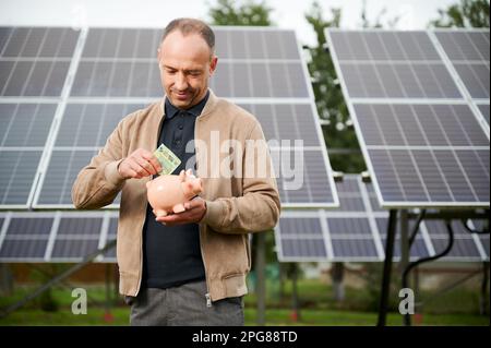 Uomo di fiducia che mette i soldi guadagnati alla banca del piggy. Il profitto di risparmio dell'adulto maschio ha ottenuto investendo nell'energia verde. Uomo in abiti casual tenendo in mano la scatola del denaro e mettere euro in esso. Foto Stock