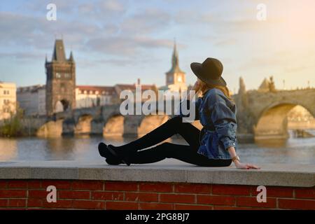 Elegante bella giovane donna con cappello nero seduto sulla riva del fiume Moldava a Praga con Ponte Carlo sullo sfondo. Messa a fuoco in primo piano. Foto Stock