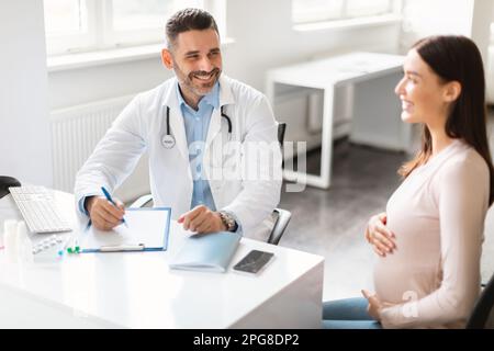 Concetto di cura prenatale. Felice giovane donna incinta seduta in clinica, avendo consulation con ginecologo maschile Foto Stock