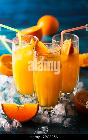 Succo d'arancia fresco in bicchieri, cubetti di ghiaccio e una fetta di frutta sul tavolo blu. Bevanda sana della vitamina. Foto Stock