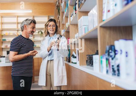 Farmacista che mostra ad un uomo un prodotto per la cura della pelle in un chimico. Operatrice sanitaria femminile che assiste un paziente in farmacia. Foto Stock