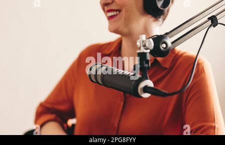 Donna felice sorridente e che guarda lontano mentre si siede dietro un microfono con cuffie. Donna allegra intervistando un ospite mentre ospita una radio dal vivo Foto Stock