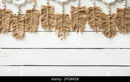 Macrame a forma di foglia o piuma marrone chiaro che pende sullo sfondo di legno bianco nella stanza di casa. Molto spazio di copia, boho sfondo. Foto Stock