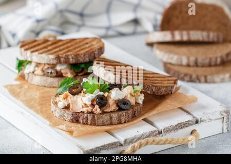 Sandwich con pesce al forno (tonno o salmone) con olive nere, formaggio Feta e prezzemolo su pane di segale. Colazione mediterranea a base di pesce. Messa a fuoco selettiva Foto Stock