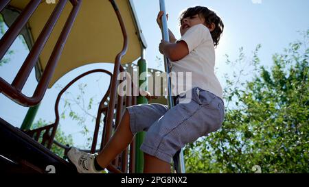 Il bambino scivola giù dal palo di metallo del parco giochi durante la bella giornata di sole al parco cittadino. Bambino attivo che scorre verso il basso con retroilluminazione a luce parassita Foto Stock