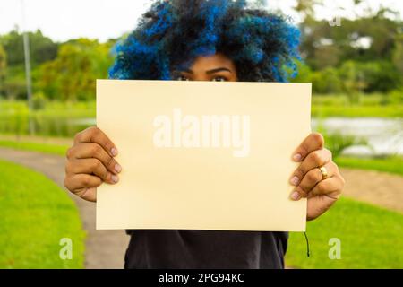 Goiania, Goias, Brasile – 21 marzo 2023: Una giovane donna, con capelli tinti di blu, il suo volto nascosto dietro un poster bianco, con un paesaggio sullo sfondo Foto Stock