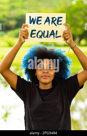 Goiania, Goias, Brasile – 21 marzo 2023: Una giovane donna, dai capelli tinti blu, con un segno con il testo: 'Siamo uguali', con un paesaggio nel bac Foto Stock