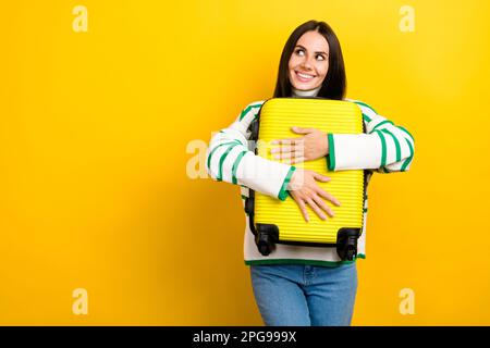Foto di carino adorabile donna indossare a strisce maglione abbracciando il bagaglio cercando spazio vuoto isolato sfondo di colore giallo Foto Stock
